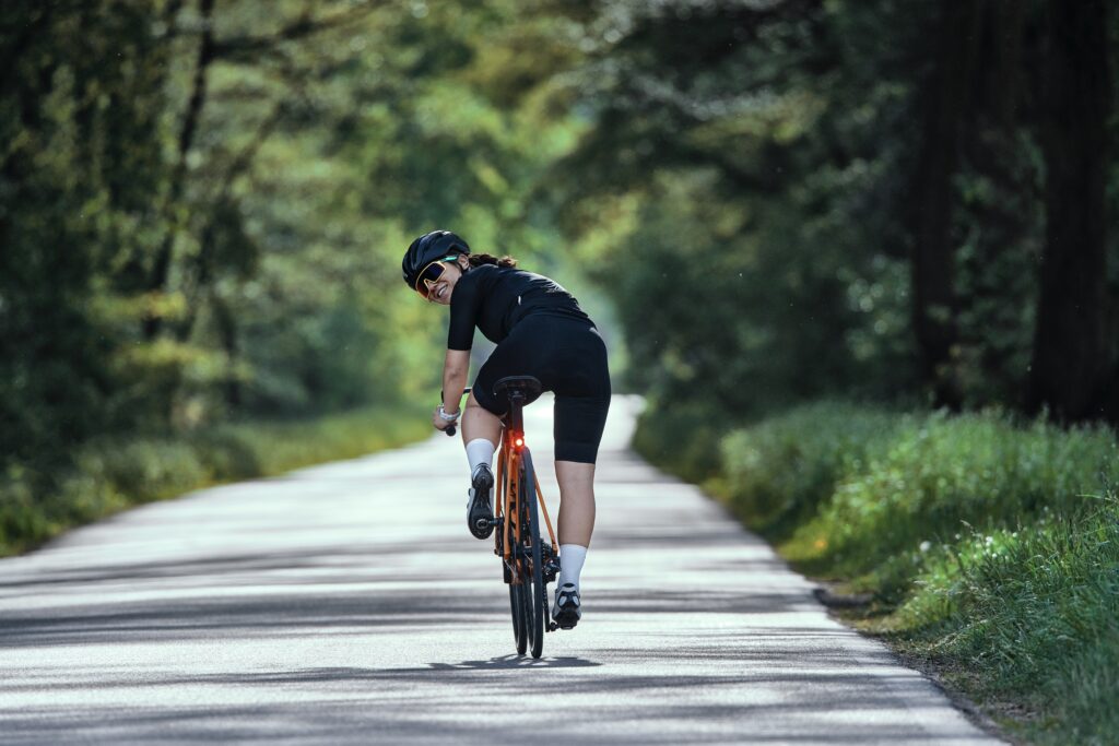 Na zdjęciu widać uśmiechniętą kobietę jadącą na rowerze szosowym drogą asfaltową przez las. Kobieta odwraca się i się uśmiecha do obiektywu