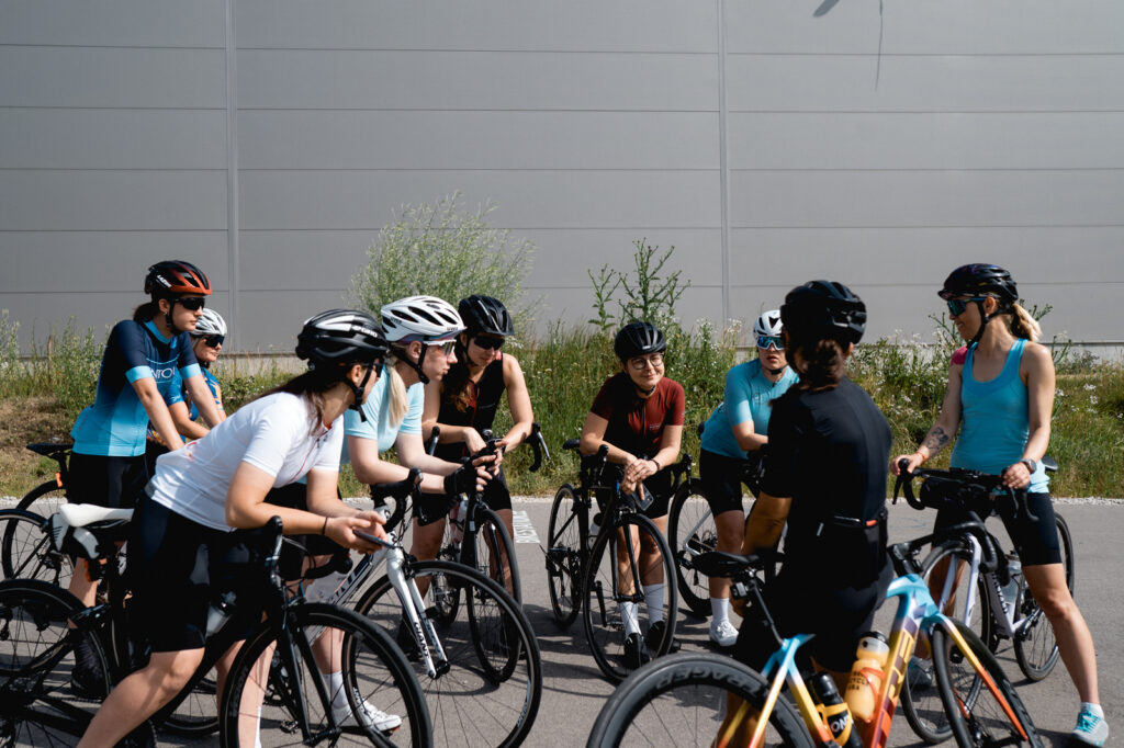 Na zdjęciu widać grupę kolarek szosowych podczas odprawy, na której organizatorka, Ministra Kolarstwa omawia zasady